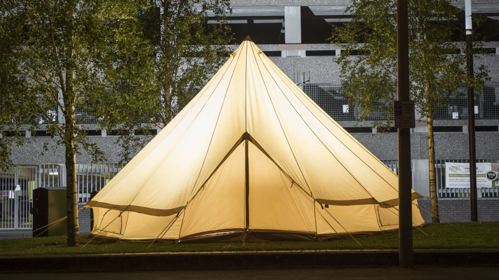 |A bell tent with a light inside stands between trees in front of a multi-storey carpark|A bell tent with a light inside stands between trees in front of a multi-storey carpark|
