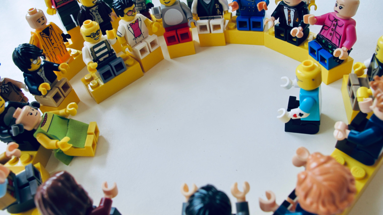 A collection of lego figurines are arranged in a circle with one lego figurine sat slightly into the circle, perhaps leading the meeting.