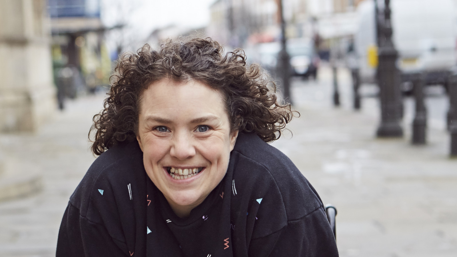 A woman with dark, curly hair and wearing dark clothes is sat in a wheelchair leaning forward towards the camera with a huge smile on her face.