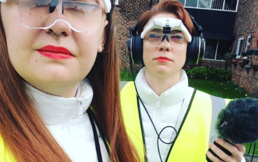 Two people stand with white and clear goggles and hi vis vests on holding a microphone.