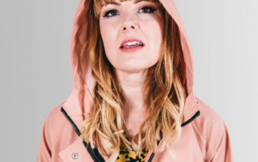 The artist Victoria Melody stands against a grey background wearing a pink coat with her hood up.