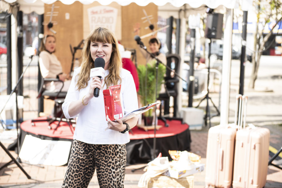 A women stands in the street with a stage behind her. She is holding a red box and a microphone. Behind her, at the stage two women wearing beige dresses and TV aerials on their heads sit looking towards the women with the microphone.