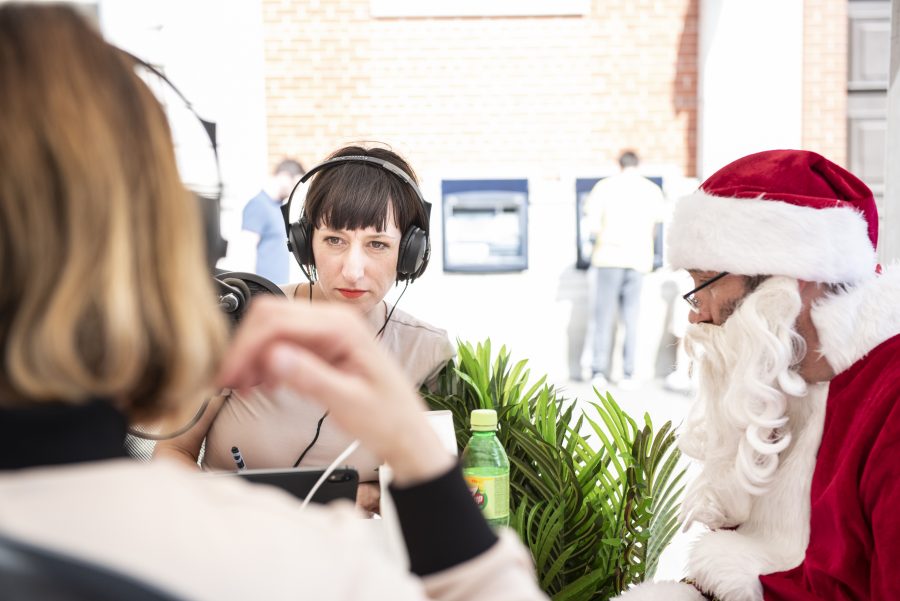 A woman in a beige dress wearing headphones looks at another woman wearing headphones. A man dressed as Santa Clause sits looking at them.