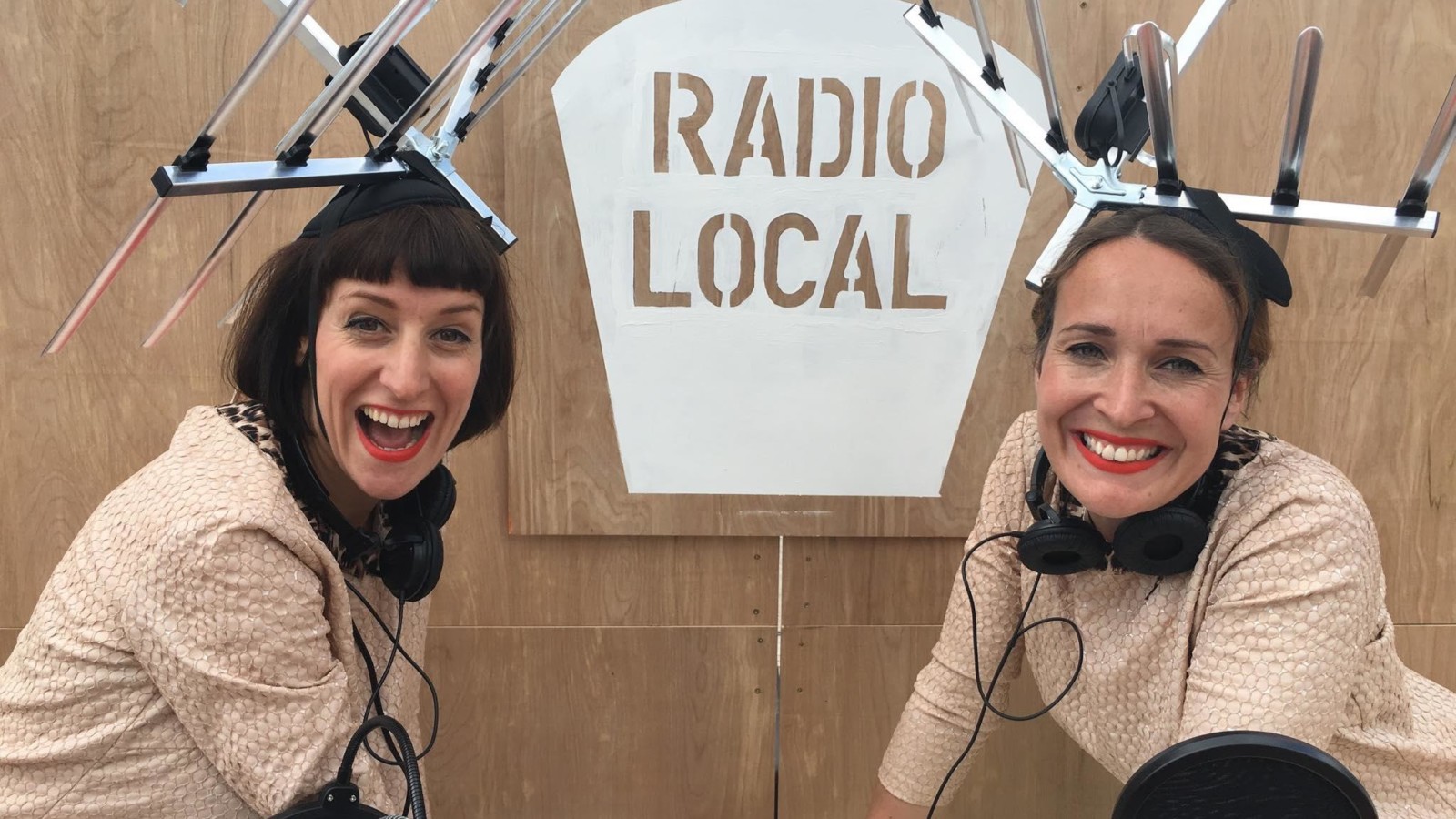 Two women wearing beige dresses, headphones and TV aerials on their heads smile in front of a wooden sign which reads ' Radio Local'